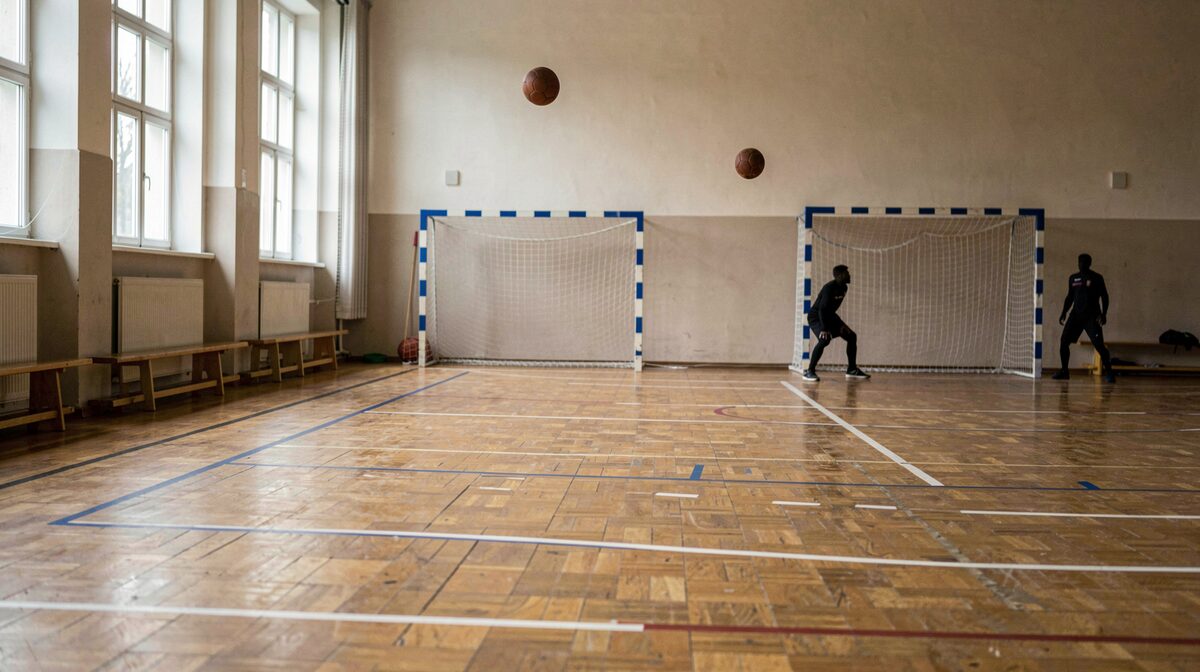 Käsipallokenttä koulun salissa Itä-Helsingissä: maalit molemmissa päissä, sinivalkoiset viivat parketilla, pallo irrallaan ilmassa heiton jälkeen ja pelaajien varjot lattiassa