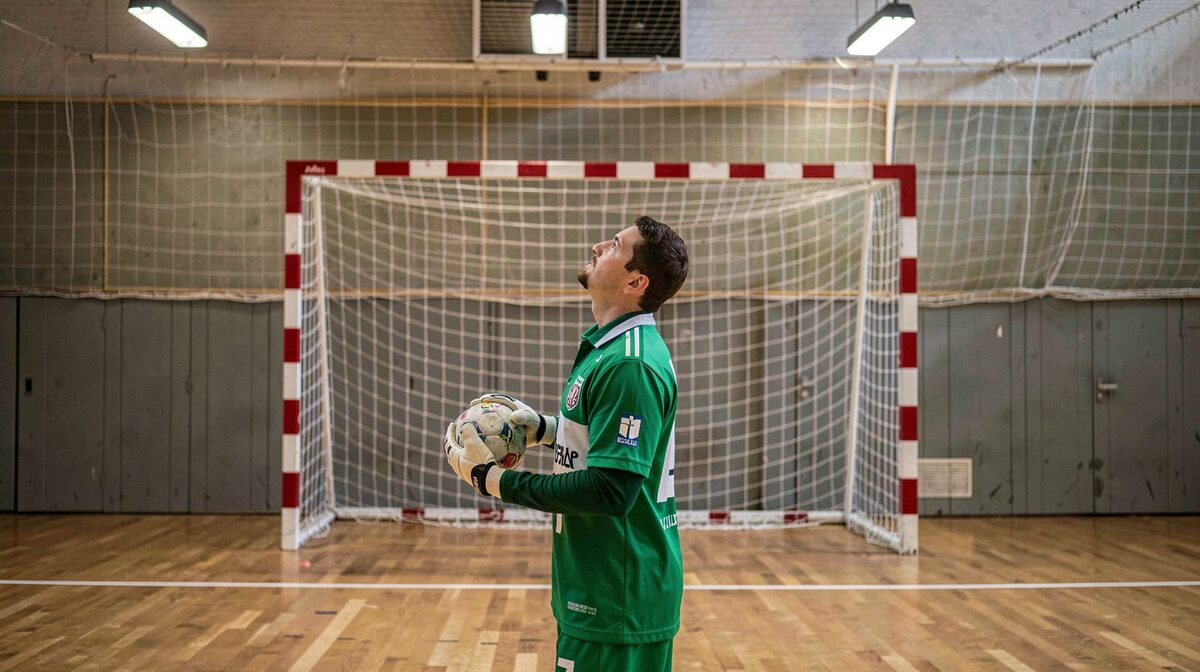 A handball goalkeeper in a green jersey standing in a goal with a net, holding the ball in one hand and looking up the court, the parquet floor and the six-metre arc in front of the goal, hall lights above, no people in the foreground